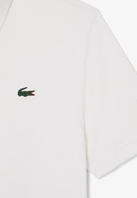 Hvid polo T-shirt lavet af bomuld, med korte ærmer og et grønt krokodille-logo på venstre bryst. Glat tekstur og klassisk kravedesign.