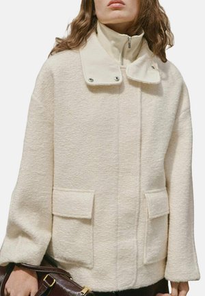 Veste de couleur crème en tissu texturé. Présente un col montant avec des pressions, deux poches avant et des manches amples.