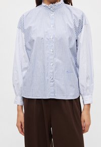 Femme portant une chemise blanche à manches longues boutonnée avec de fines rayures verticales bleues et un pantalon marron sur un fond uni.