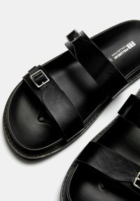Sandales en cuir noir avec deux larges sangles munies de boucles en argent, une doublure intérieure texturée et une semelle plate avec des détails contrastants.