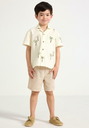Smilende dreng iført en cremefarvet kortærmet skjorte med grønne palmebladtryk, beige shorts og brune snørebåndssko, stående mod en hvid baggrund.