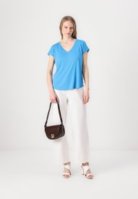 Modrá krátkorpová V-krčná blúzka, biele široké nohavice, hnedá crossbody taška a biele šľapky na podpätku, zamerané na čisté línie a jasné farby.