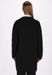 Langer schwarzer Cardigan aus weichem Material, mit lockerer Passform, abgesenkten Schultern und einem dezenten Logodetail am hinteren Halsauschnitt.