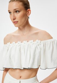 Crop top bianco off-the-shoulder con design arricciato, applicazioni floreali e bordi neri. Tessuto liscio con vestibilità rilassata e dettagli texturizzati.