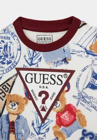 Sweatshirt blanche avec col côtelé bordeaux, ornée d'illustrations d'ours bleus et bruns, divers logos, et une étiquette triangulaire "GUESS".