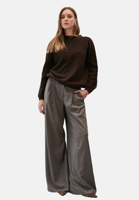 Brązowa bluza z ściągaczami, zestawiona z szarymi, wysokim stanie, plisowanymi spodniami o szerokich nogawkach. Model stoi z rękami w kieszeniach.