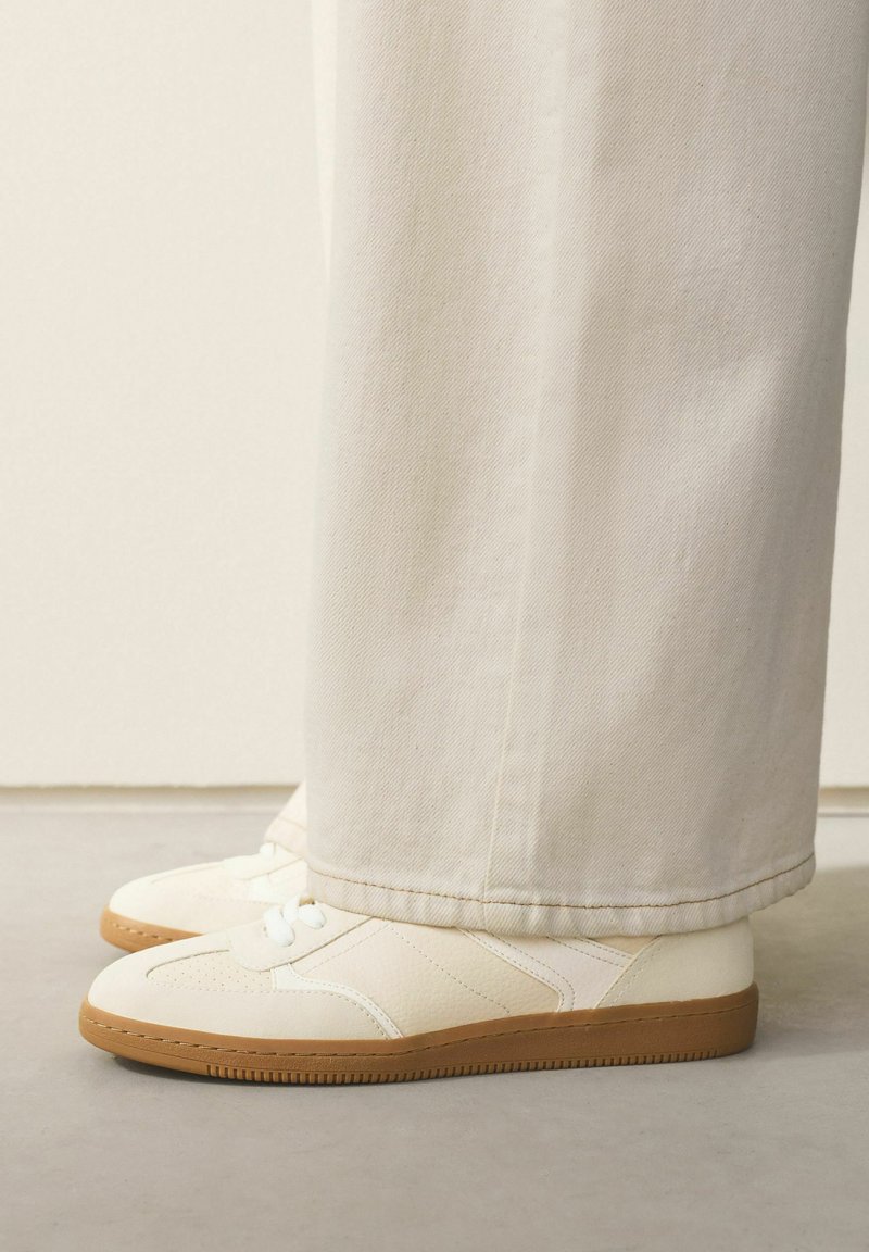 Balti batai su tekstūruotais intarpais ir guminiais padais, derinami su kreminės spalvos plačiomis kelnėmis, matoma tik apatinė dalis.