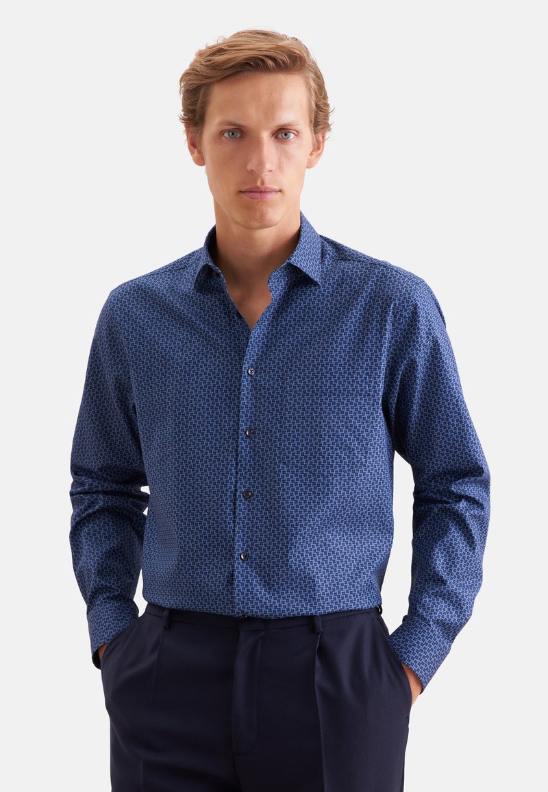 Homme aux cheveux clairs portant une chemise bleue à manches longues à motifs et un pantalon foncé, debout les mains dans les poches sur fond blanc.