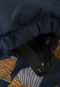 Marineblå tygkåpa med samlad elastik, prydd med mörkgröna justerbara remmar. Bakgrunden visar ett geometriskt mönster i gult och blått.