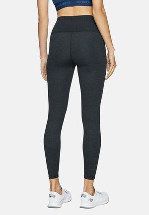 Femme portant un legging noir taille haute, un soutien-gorge de sport bleu marine et des baskets blanches New Balance, debout, dos tourné sur un fond uni.