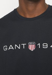 Camiseta de algodón negra con el texto blanco "GANT" y un logotipo en forma de escudo con franjas rojas y blancas y tres estrellas, diseño de cuello redondo.