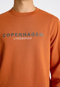 Oranje sweatshirt met een ronde hals, gemaakt van zacht materiaal. Bevat bedrukte tekst: "KOPENHAGEN" in donkergrijs en "LINDBERGH" in het wit.