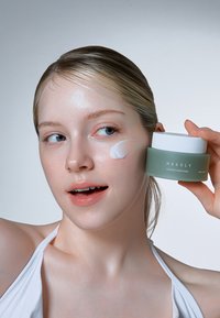 Crema reparadora Cicachild en un frasco verde mate con tapa blanca, sostenido junto al rostro de una modelo. Crema aplicada en su mejilla, textura suave.
