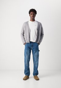 Grå strikket cardigan over en hvit T-skjorte, kombinert med blå cargo jeans og brune slip-on sko. Enkel design, avslappet passform.