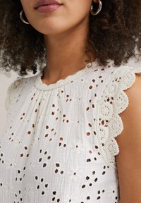 Witte blouse van lichtgewicht stof, met geborduurde bloemenpatronen, geschulpte kanten rand en subtiele plooien bij de halslijn.