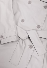 Trenci gri deschis cu nasturi cu două rânduri, curea de talie legată într-un nod și detaliu de manșetă cu nasturi.