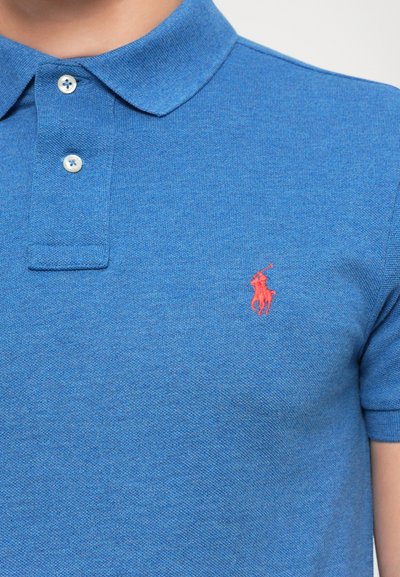 Zils īsās piedurkņu polo krekls ar divām baltām pogām un mazu sarkanu izšūtu polo spēlētāju, kas jāja uz zirga, uz krūtīm.