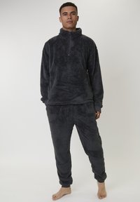 Jeune homme se tenant pieds nus, portant un sweat à capuche gris foncé en peluche et un pantalon assorti, devant un fond clair.