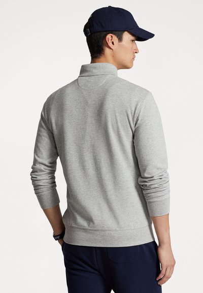 Grijze sweatshirt met een hoge kraag, geribde manchetten en zoom. Het model draagt een marineblauwe pet. Het materiaal lijkt zacht met een gladde textuur.
