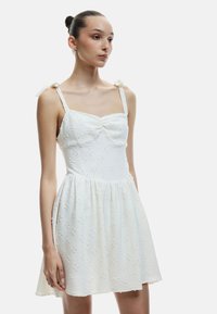 Vestido blanco con un corsé ajustado, falda fruncida y tela texturada con flores. Presenta tirantes finos y lazos de tela en los hombros.