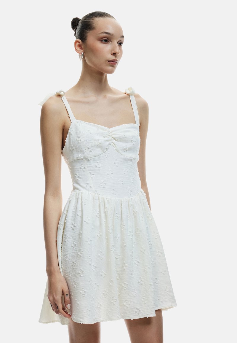 Vestido blanco con un corsé ajustado, falda fruncida y tela texturada con flores. Presenta tirantes finos y lazos de tela en los hombros.