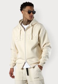 Veste à capuche zippée beige avec logo brodé "ESTM", capuche avec cordon de serrage, poches latérales et poignets côtelés, assortie à un pantalon beige.