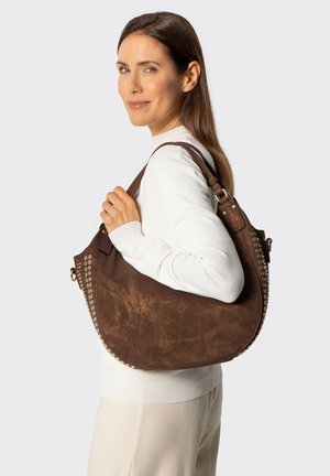 Frau im weißen Pullover hält große braune Lederschultertasche mit Metallnieten und Wittchen-Logo und lächelt seitlich.