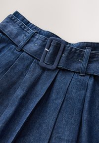 Spijkerrok in donkerblauw met een brede tailleband voorzien van een rechthoekige gesp en plooien die vanaf de taille vloeien. Soevele textuur.