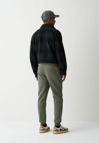 Jachetă neagră din fleece, pantaloni de trening verzi măslinii, șapcă gri și adidași bej. Ăsta este un ansamblu cu un croi lejer și buzunare pe pantaloni.