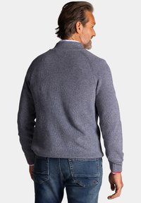 Grijze gebreide trui met een textuurafwerking, voorzien van een kraag en geribde manchetten. De man draagt een spijkerbroek in het blauw met een rode accent op de manchetten.