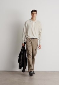 Ανοιχτό μπεζ μακρυμάνικο polo μπλουζάκι συνδυασμένο με καμηλό cargo παντελόνι. Το μοντέλο κρατάει μια μαύρη δερμάτινη ζακέτα. Ήπια ύφανση και χαλαρός στυλ.