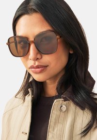 Lunettes de soleil carrées surdimensionnées marron avec une monture en écaille et des verres marron. Portées avec une veste beige dotée d'un col en velours côtelé.