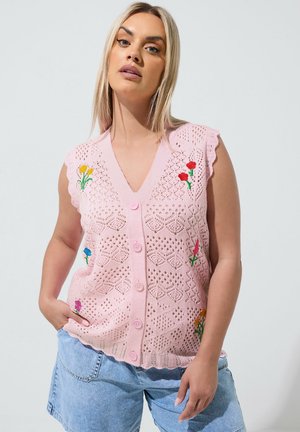 Kvinde med blond hår iført en pink ærmeløs strik top med blomsterbroderi og blå denimshorts, stående med den ene hånd i lommen.