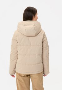 Beige donsjack met capuchon, voorzien van een doorgestikt patroon en zichtbare stiksels. De stof lijkt glad en licht glanzend te zijn.