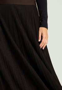 Falda negra plisada hecha de tela brillante, con una amplia banda marrón en la cintura. Primer plano muestra una mano tocando la textura de la tela.