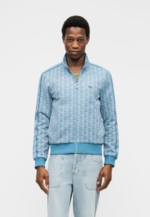 Jeune homme portant une veste zippée à motifs bleu clair et un jean délavé, debout devant un fond blanc uni.
