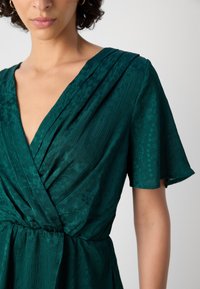 Femme portant une robe portefeuille texturée vert profond avec des manches courtes volantées et des boucles d'oreilles créoles dorées sur un fond uni.