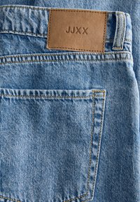 Κοντινή λήψη της πίσω τσέπης ενός μπλε τζιν με καφέ δερμάτινο σημείο με την επιγραφή "JJXX" ραμμένη πάνω από τη μέση.