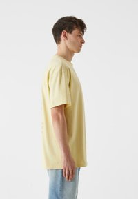 Trumpas rankovės, apvalaus kaklo marškinėliai pastelinių geltonų spalvų medvilnės audinio. Minkšta tekstūra, laisvas siluetas, minimalus raštas ant nugaros.