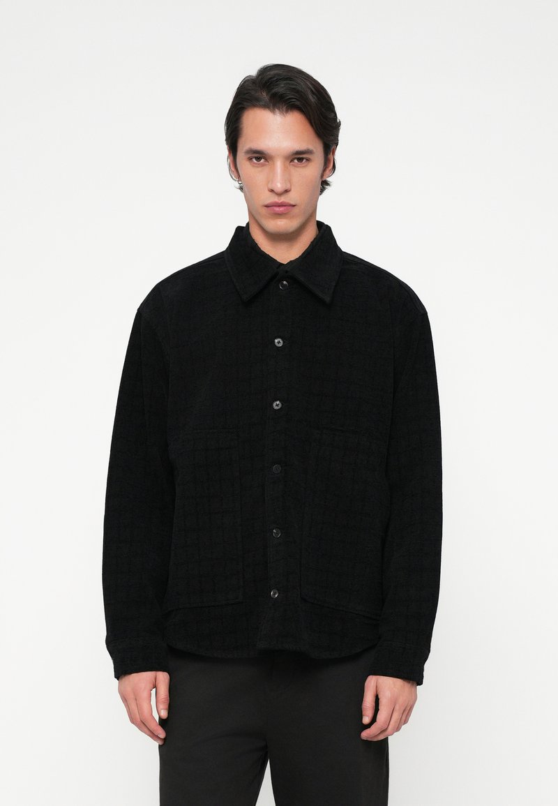 Άνδρας που φοράει ένα μαύρο υφασμάτινο πουκάμισο με κουμπιά και γιακά, με μεγάλες μπροστινές τσέπες, που στέκεται μπροστά σε λευκό φόντο.