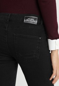 Svarta denimjeans med en medelhög design, som har en bakficka och en etikett vid midjan. Tyget verkar slätt med l ätt struktur.