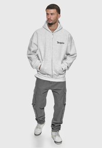 Haut à zip gris avec de grandes poches et un logo sur la poitrine, associé à un pantalon cargo gris et des baskets blanches. Texture de tissu lisse.