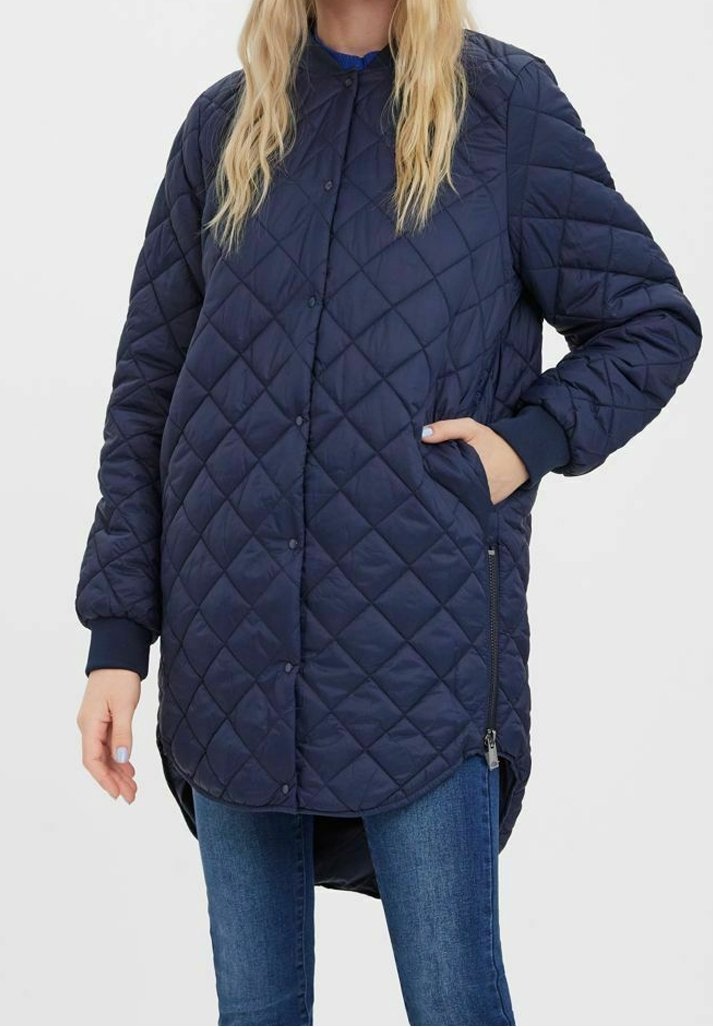 Femme portant une veste matelassée bleu marine avec boutons pression, zips latéraux, poignets côtelés et la main dans la poche, associée à un jean bleu.