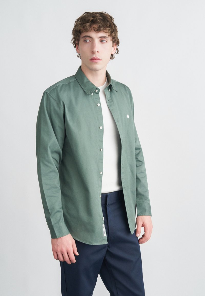 Πράσινο πουκάμισο με κουμπιά από μαλακό βαμβάκι με γιακά. Διαθέτει μικρό λογότυπο στο στήθος και μακριά μανίκια, σε συνδυασμό με σκούρο μπλε παντελόνι.