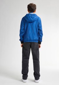 Jachetă cu glugă albastră, din material rezistent la vânt, cu un design texturat; mansete și tiv negru cu nervuri. Asortată cu pantaloni cargo negri și pantofi sport albi. Vedere din spate.