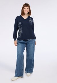 Suéter de punto azul marino con escote en V y bordado floral blanco en el lado izquierdo, combinado con jeans de pierna ancha y zapatos blancos.