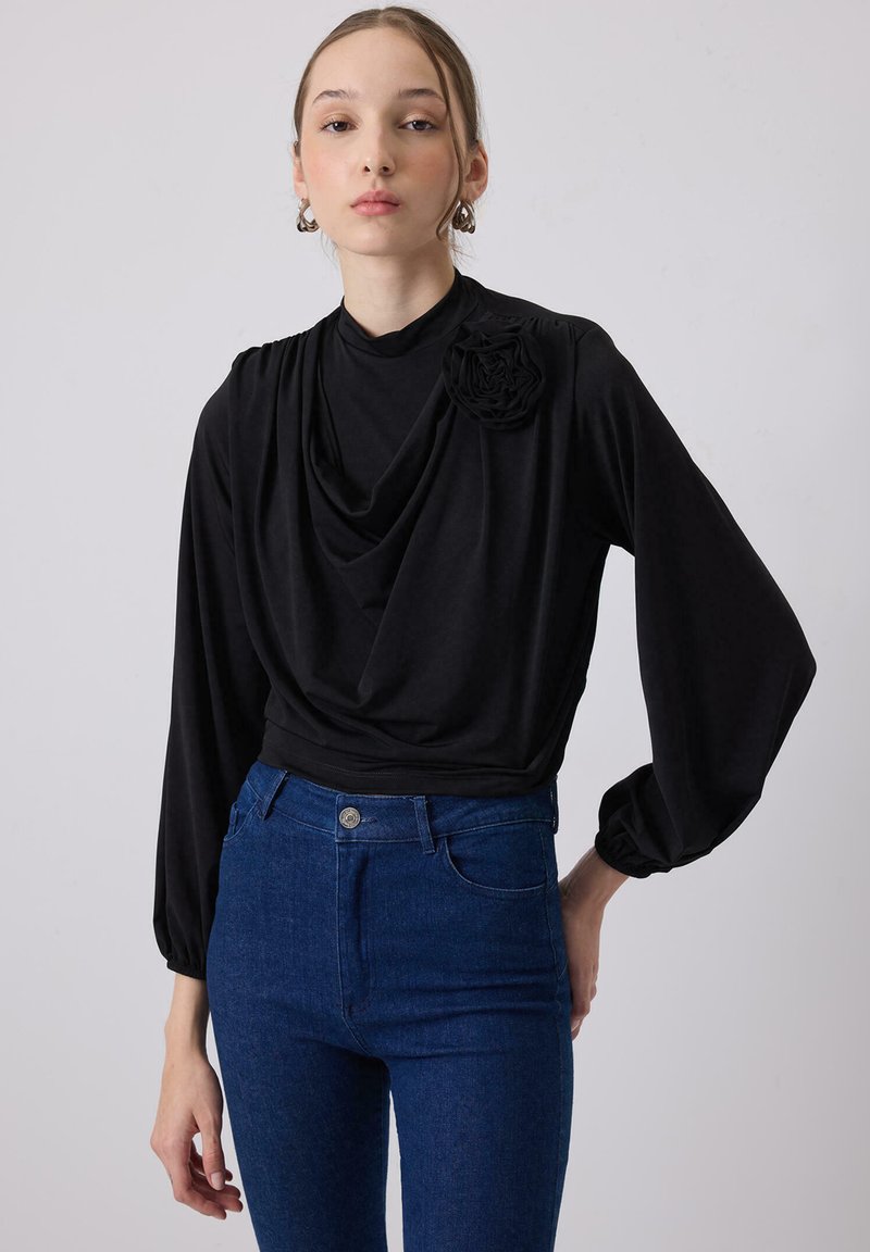 Maglietta nera a maniche lunghe con scollo drappeggiato e dettaglio di rosa sulla spalla, abbinata a jeans a vita alta blu scuro.
