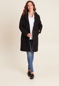 Zwarte wollen jas met een reverskraag, vijf knopen en zijzakken, gecombineerd met een wit shirt en blauwe spijkerbroek. Gladde textuur, knielang.