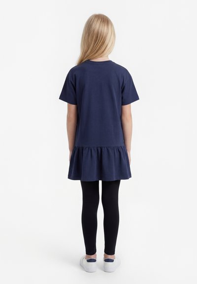 Παιδί με μακριά ξανθά μαλλιά, που φοράει ένα φόρεμα σε σκούρο μπλε χρώμα με κοντά μανίκια και βολάν στο τελείωμα, μαύρο κολάν και λευκά αθλητικά παπούτσια, γυρισμένο ανάποδα.