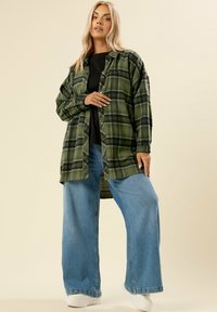 Camicia di flanella a quadri verdi con accenti neri, chiusura a bottoni, abbinata a jeans in denim blu larghi e sneakers bianche. Texture liscia.